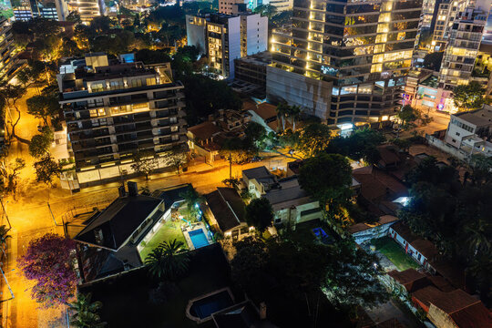 Aerial view of the illuminated Las Lomas neighborhood featuring modern high-rise buildings, residential houses with swimming pools, and tree-lined streets at night in Asuncion, Asuncion, Paraguay.