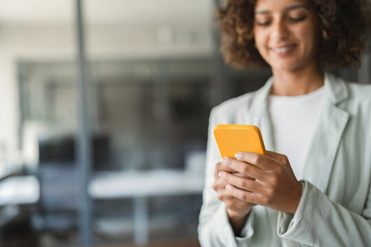 Sitting at desk in office, indian businesswoman using smartphone, smiling, working beside open laptop. Young hispanic business woman, accountant, coordinates tasks at workplace daily. Copy space