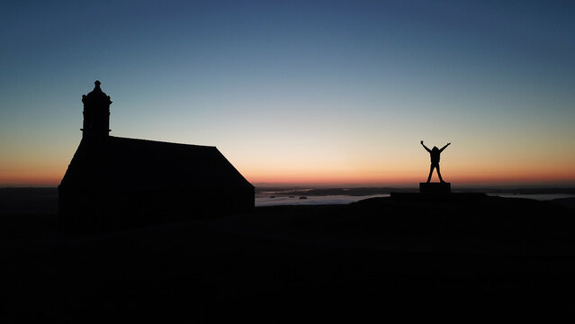 Aerial view of Mont Saint-Michel de Brasparts chapel silhouette with a person standing with arms raised against a colorful sunrise sky in Saint-Rivoal, Bretagne, France.