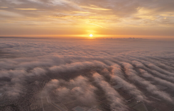 Aerial view of a vast landscape blanketed by a thick layer of low-lying clouds at sunset, with the sun glowing on the horizon above the fields and roads of Ginosa, Apulia, Italy.