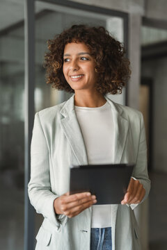 Vertical portrait of indian businesswoman standing in office, using tablet, smiling, working on growth strategy. Young hispanic business woman, financial analyst, plans ahead at workplace daily.