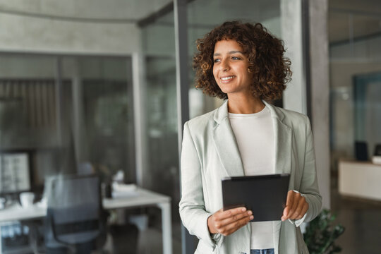 Standing in office corridor, curly haired latina businesswoman using tablet, smiling and glancing away. Young arab business woman, sales manager, checks client email at corporate workplace.