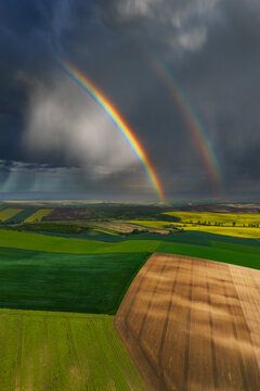 Aerial view of a double rainbow arching over vibrant agricultural fields with green and brown patterns under a dramatic stormy sky in Nasedlovice, South Moravian Region, Czechia.