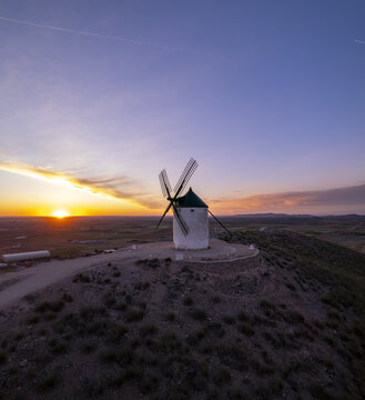 Aerial view of Molinos de Viento de Consuegra standing on a hilltop during a vibrant sunset with golden light illuminating the surrounding landscape in Consuegra, Castilla-La Mancha, Spain.
