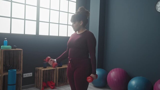 Woman lifting a red dumbbell with a bicep curl in studio gym with exercise balls and wooden crates; steady determination.