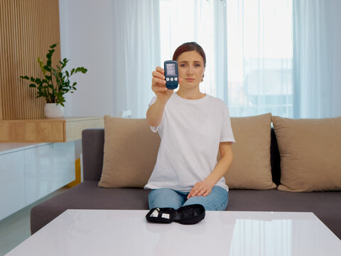 A woman displays a glucometer reading of 200 mg dL while seated in her living room. This reading indicates a high glucose level that needs attention.