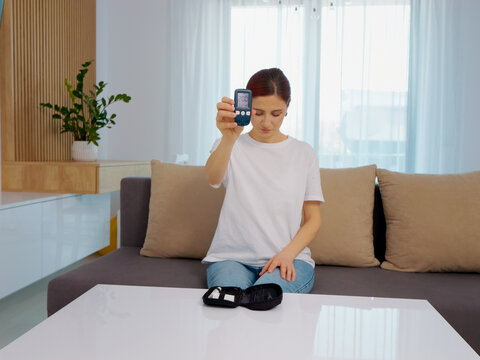 A woman holds a glucometer displaying a reading of 200 mg dL. This shows a high blood sugar level. She looks concerned while sitting on a couch in her home.