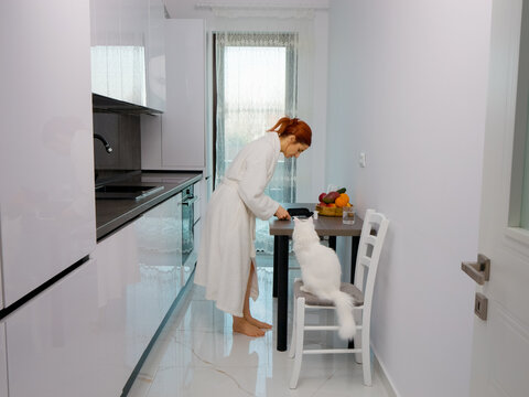 A woman measures her blood glucose level in the kitchen. She focuses on the glucometer while standing by a table next to some fresh fruit and her pet cat.