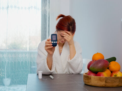 A woman measures her blood sugar level in the kitchen. The glucometer shows a high reading of 141 mg dL. She reflects on her health and dietary choices.