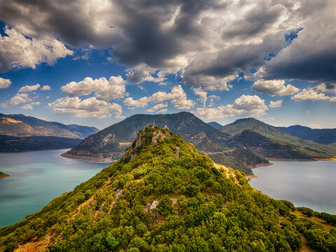 Aerial view of Techniti Limni Arachthou surrounded by lush green hills and calm blue waters under a dramatic cloudy sky in Arta, Greece.