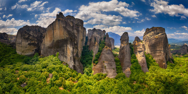 Aerial view of Meteora rock formations surrounded by lush green forests under a bright blue sky with scattered clouds in Trikala, Greece.