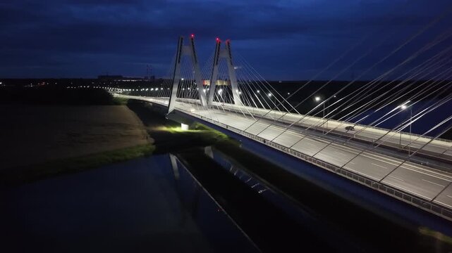 Vehicles crossing over modern cable-stayed bridge at night illuminated by streetlights. Cars riding on a dark road. Autos drive at evening highway. Aerial view