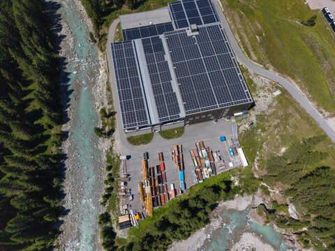 Aerial view of the Upper Engadine Wastewater Treatment Plant with rooftop solar panels, a storage yard, and the adjacent river in S-chanf, Grisons, Switzerland.