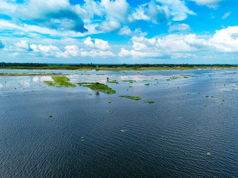 Aerial view of the flooded landscape in Kushtia, featuring lush green vegetation, calm water, and a bright blue sky with white clouds in Kushtia, Khulna Division, Bangladesh.