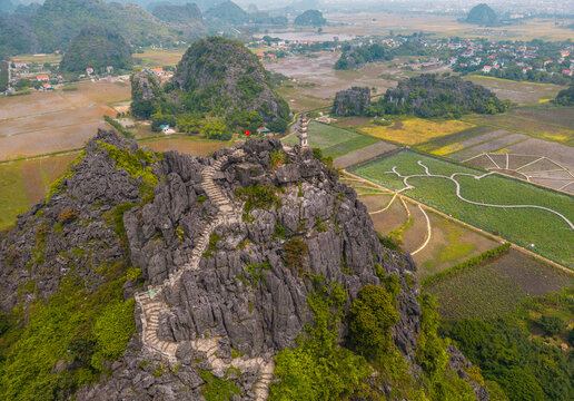 Aerial view of Mua Cave with its iconic stone staircase winding up the karst mountain, surrounded by lush green fields and limestone formations in Nam Hoa Lu, Ninh Binh, Vietnam.