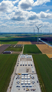 Aerial view of a large battery energy storage system facility surrounded by wind turbines and agricultural fields in the rural landscape of Dronten, Flevoland, Netherlands.