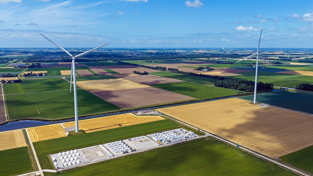 Aerial view of wind turbines standing amidst vast agricultural fields and a storage facility under a clear blue sky in Dronten, Flevoland, Netherlands.