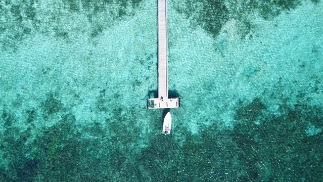Aerial view of a wooden pier extending into clear turquoise water with a small boat docked at the end over a vibrant coral reef in Indonesia.