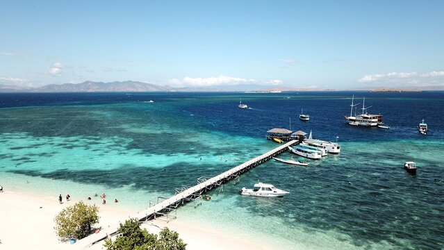Aerial view of a wooden pier extending into turquoise waters with boats moored at the end, a white sand beach with people swimming, and distant Kanawa islands in Indonesia.