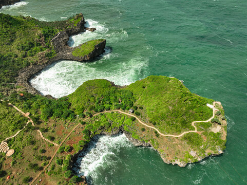 Aerial view of Pantai Kesirat with rugged green cliffs, winding paths, and turquoise ocean waves crashing against the rocky coastline in Panggang, Special Region of Yogyakarta, Indonesia.