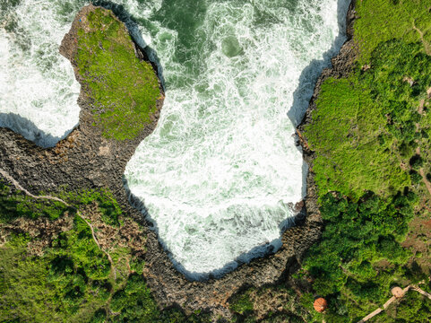 Aerial view of Pantai Kesirat showing dramatic waves crashing against rugged limestone cliffs surrounded by lush green vegetation in Panggang, Special Region of Yogyakarta, Indonesia.