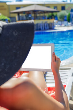 A woman in a black sun hat relaxes on a deck chair near a swimming pool while holding a digital tablet with a blank white screen.