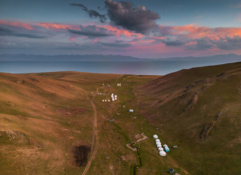 Aerial view of Song-Kul lake surrounded by rolling hills and a traditional yurt camp under a vibrant sunset sky in Naryn Region, Kyrgyzstan.