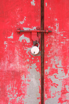 Close-up of a weathered red metal door secured with a padlock, featuring peeling paint and a rustic textured surface