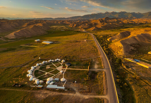 Aerial view of a yurt camp on grassy plains near a road, surrounded by rolling hills and mountains under the warm glow of the setting sun in Naryn, Naryn Region, Kyrgyzstan.