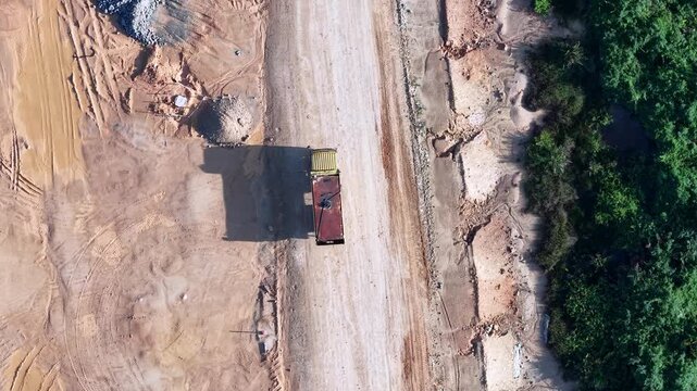Aerial view shows dirt road through construction site. Red dump truck travels along the unpaved path