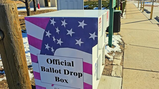 Official ballot drop box decorated with American flag stars and stripes stands beside a snowy sidewalk, offering secure mail-in voting during local election season in bright winter sunlight.
