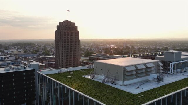 Green rooftop garden spreads beside mechanical penthouse and rows of HVAC louvers, Art Deco tower rises above hazy Allentown skyline at golden sunset in Pennsylvania. Aerial drone flyover shot