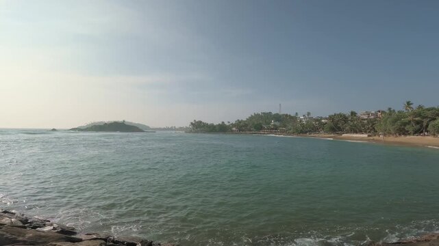 A slow rightward pan reveals turquoise water, a palm-lined sandy beach and lush tropical coastline in Mirissa, Sri Lanka, with a small offshore islet beneath a hazy blue sky.