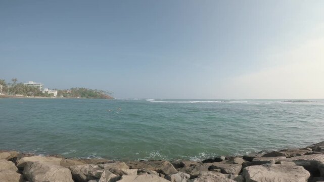 A slow rightward pan from foreground boulders opens onto rippled turquoise water, a distant tree-covered headland and bright hazy horizon on the Mirissa coast of Sri Lanka.