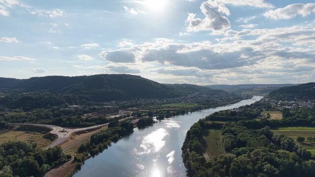Drone aerial panorama of the Moselle River in Konz, Germany, featuring calm water reflecting the evening sun, forested riverbanks and rolling hills beneath dramatic clouds. Scenic summer landscape wit