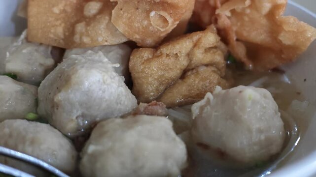 Close up slow motion shot of traditional Indonesian bakso meatball soup served with crispy wontons and fried tofu in a white ceramic bowl.
