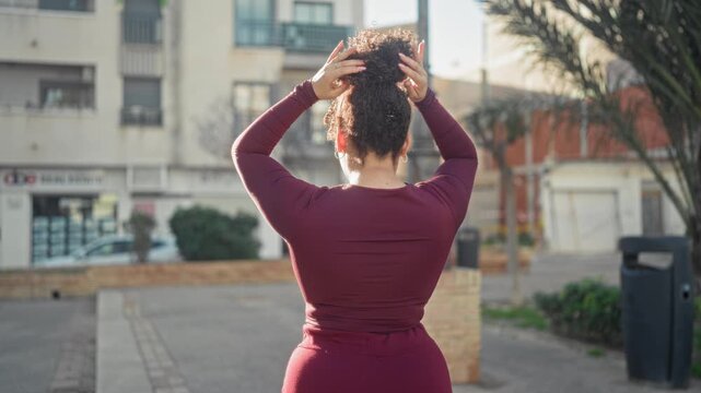 Woman tying hair on street in burgundy top and sweatpants, hands raised and smiling with lamppost and building in view; quiet confidence.