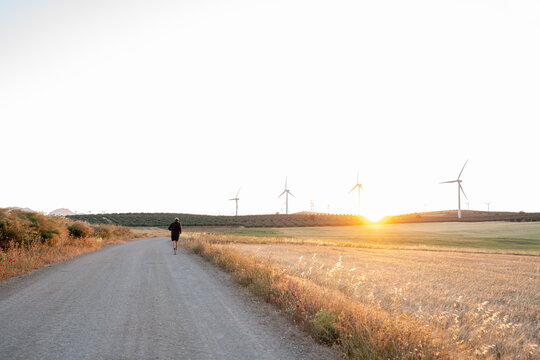 Person running on rural road at sunset with wind turbines
