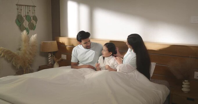 Indian man woman sitting on cozy bed do tickling to girl talk speak show love care enjoy day living room home. Young adult mom dad male female play kid child spend family time have fun joy house place