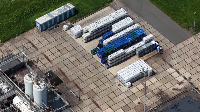 High angle perspective showing a logistics yard at a chemical plant, with rows of white and blue intermediate bulk containers neatly stacked next to large industrial storage tanks and silos