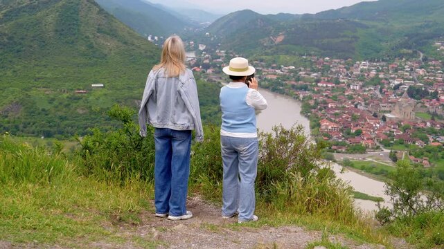 Two female tourists standing on a scenic viewpoint overlooking the ancient city of mtskheta in georgia, capturing the beautiful landscape and the confluence of the aragvi and mtkvari rivers