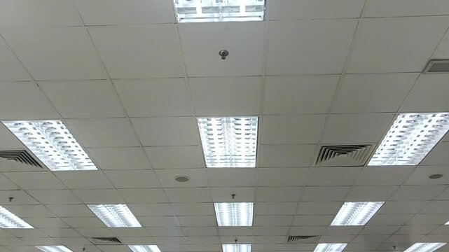  view of a commercial office ceiling featuring multiple recessed fluorescent LED light fixtures, suspended ceiling tiles, air conditioning ventilation grilles, and mounted display monitors. The interi