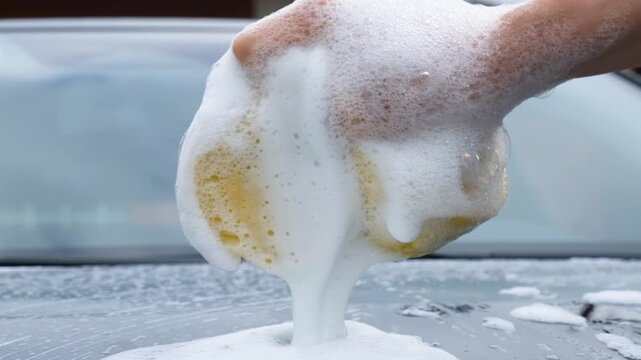 Close-up of a hand squeezing a soapy sponge over a car, releasing thick foam during vehicle washing, highlighting automotive detailing, cleaning, and maintenance.