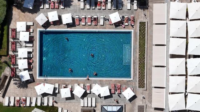 Aerial view of a rectangular swimming pool with people swimming, surrounded by rows of lounge chairs and white umbrellas in Cascais, Lisboa, Portugal.