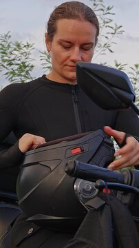 Vertical video. Female Motorcyclist Adjusts a Black Full Face Helmet Beside a Touring Motorcycle, With Coastal Hills and Sea Behind, Demonstrating Rider Preparation, Motorcycle Safety and Injury Preve