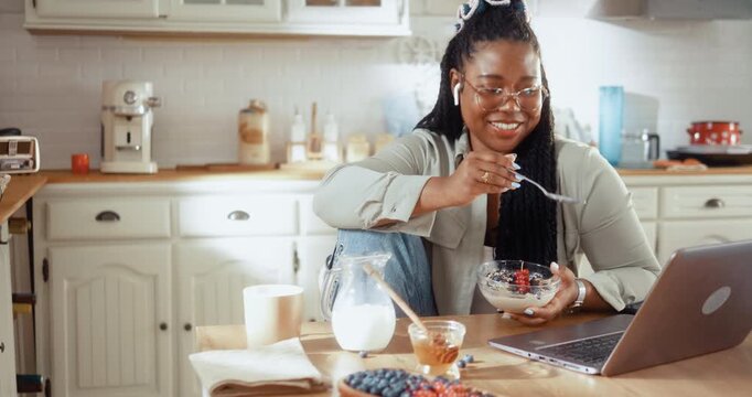 Young Black woman enjoys a healthy breakfast while having a video call on her laptop in the kitchen at home.