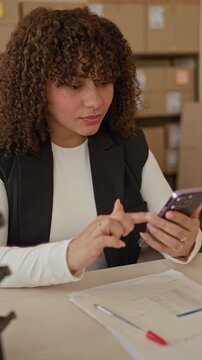 Woman holding smartphone and tapping with bare hands at a desk in a building with stacked cardboard parcels; small business focus.
