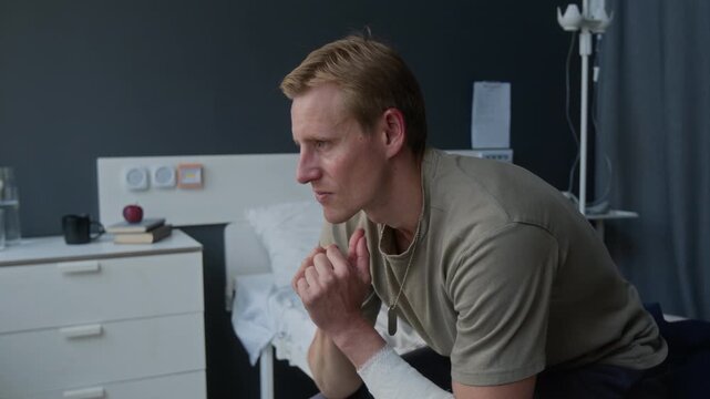 Portrait of young adult Caucasian male veteran with bandaged arm sitting on hospital bed, looking at camera and turning away. Footage for PTSD awareness and mental health campaigns