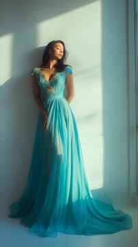 Elegant woman in a flowing turquoise evening gown stands gracefully by a sunlit window, bathed in soft natural light. Fashionable and sophisticated, she exudes timeless beauty and poise