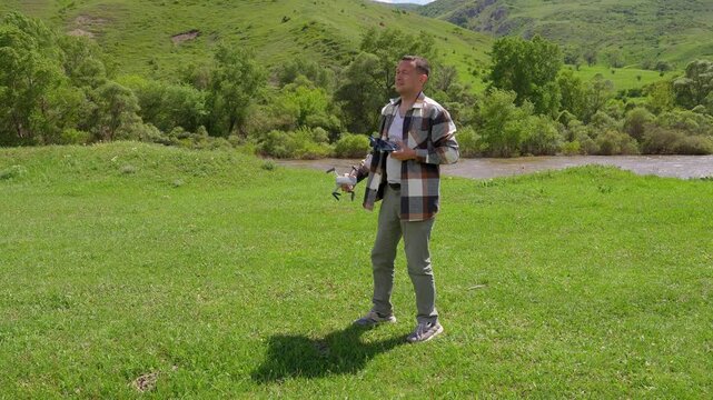 Young man in plaid shirt flying drone, landing quadcopter out of hands while standing on scenic green meadow with hills and river in background on sunny day